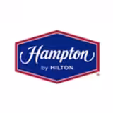 The image shows the Hampton by Hilton logo, featuring the word Hampton in white script on a blue, hexagonal background with a red border, and by Hilton in smaller text underneath.